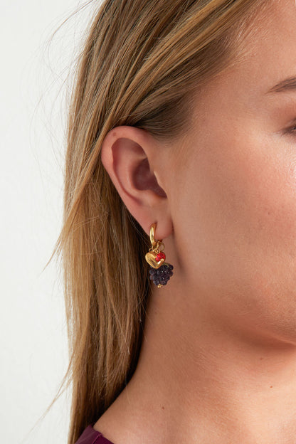 Close-up of a woman wearing gold-tone hoop earring with purple grape cluster bead, gold heart charm, and colorful crystals
