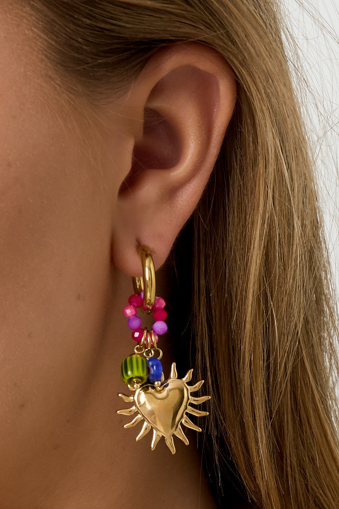 Side view close-up of a woman’s ear wearing a gold sunburst heart hoop earring with vibrant pink and green beads