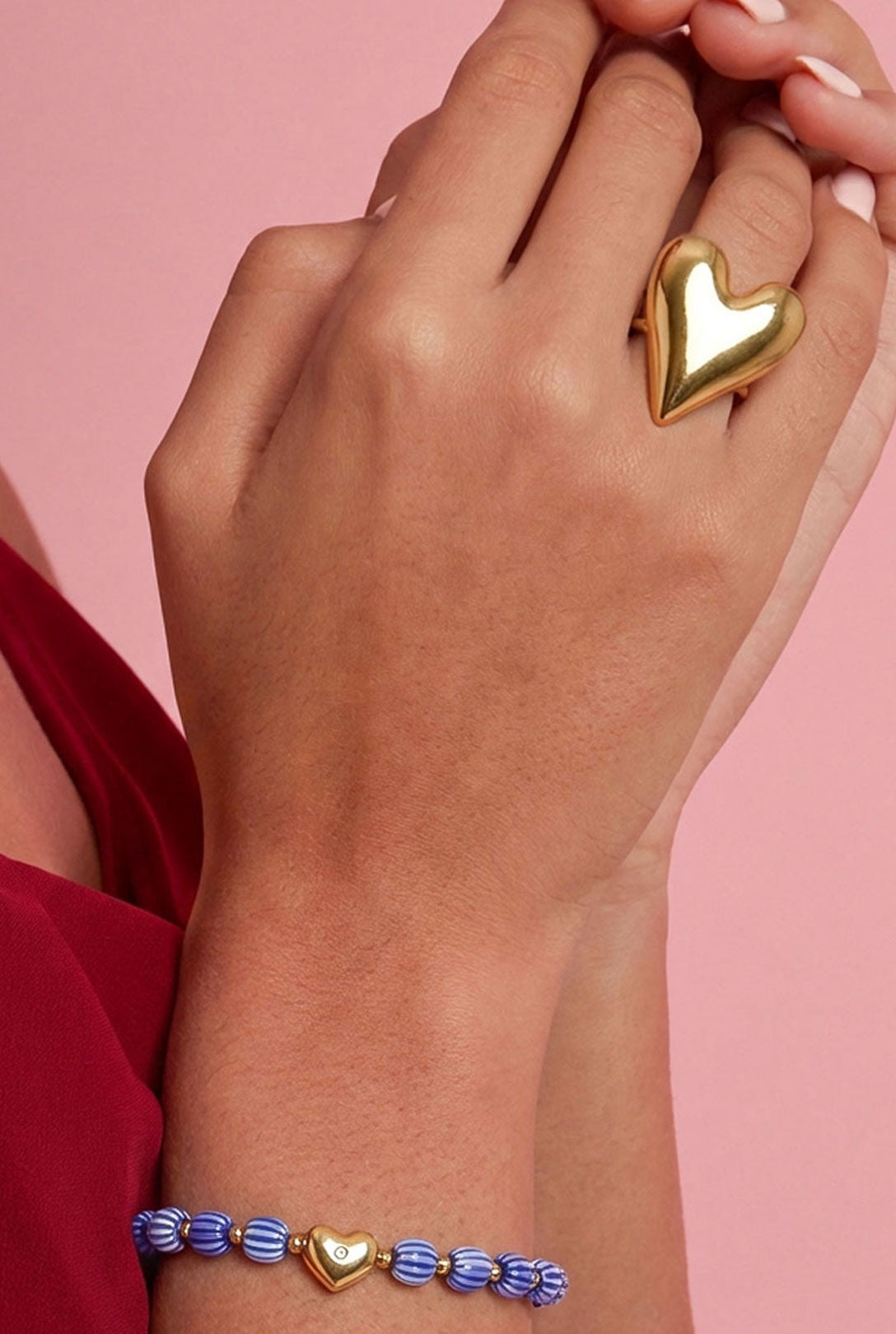 Woman’s hand wearing a large polished gold heart statement ring alongside a blue beaded bracelet with gold heart charm, against a pink background