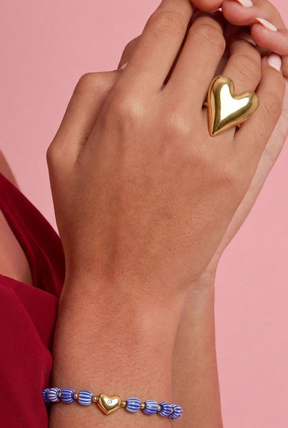 Woman’s hand wearing a large polished gold heart statement ring alongside a blue beaded bracelet with gold heart charm, against a pink background