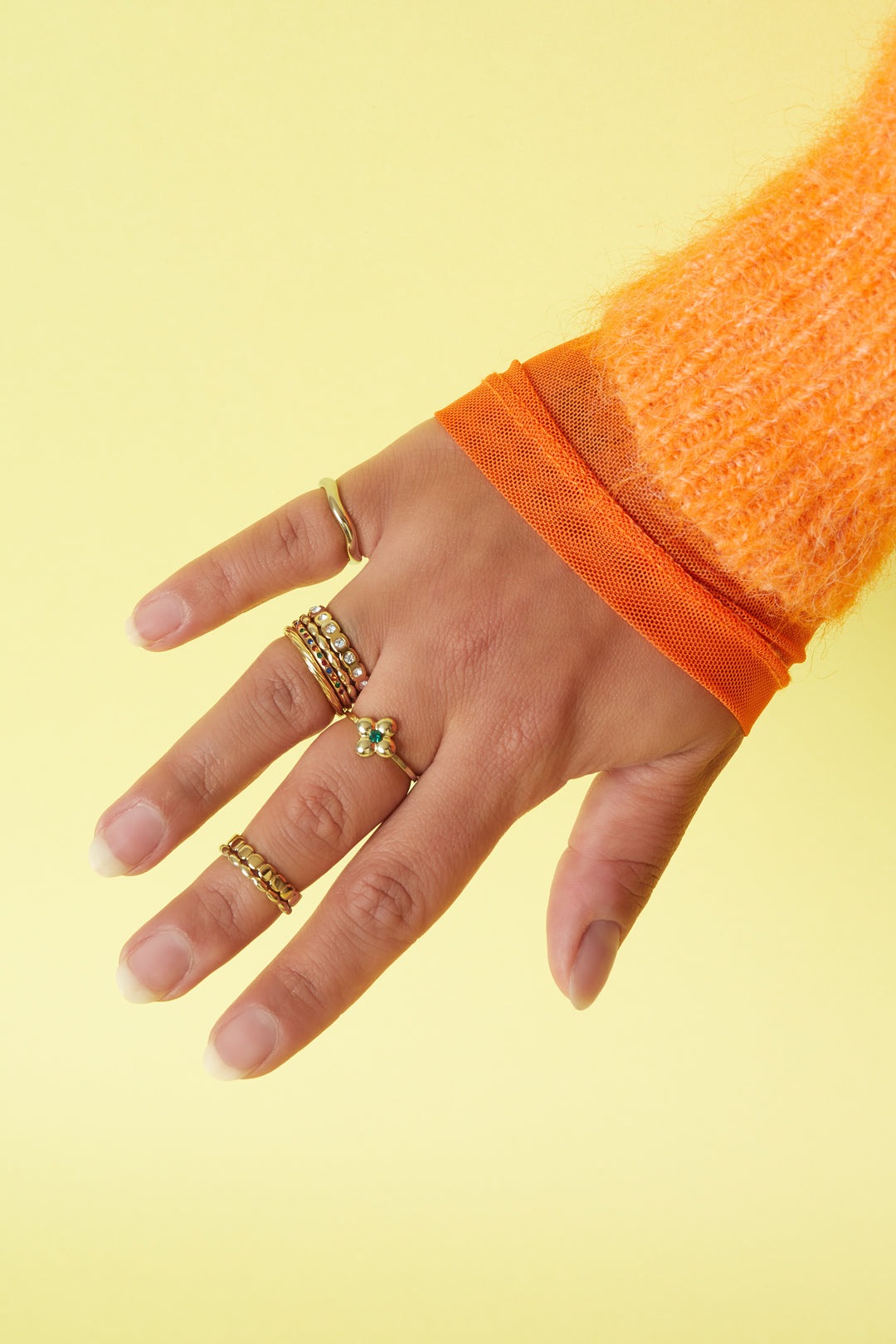 Hand wearing multiple gold rings including colorful stone ring, styled with orange sweater sleeve against yellow background