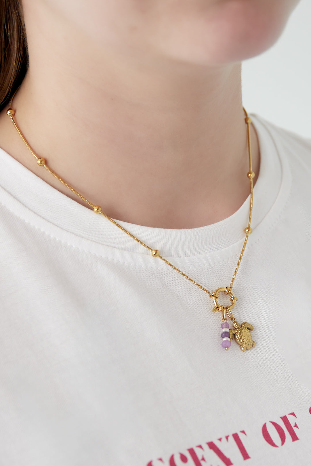 Close-up of woman wearing the gold charm necklace featuring turtle and purple bead charms against a white shirt