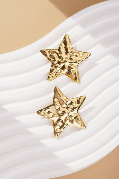 Cosmic Glitch gold star stud earrings with a textured, hammered finish, displayed on a wavy white surface.