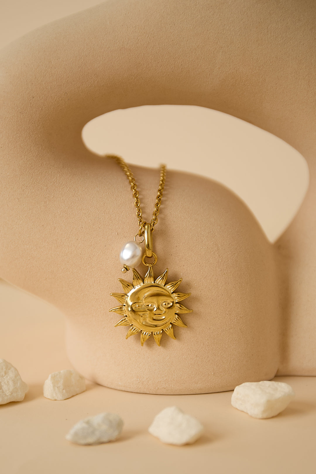 Close-up of gold sun face pendant and pearl charm necklace displayed against a soft beige sculptural backdrop with scattered white stones