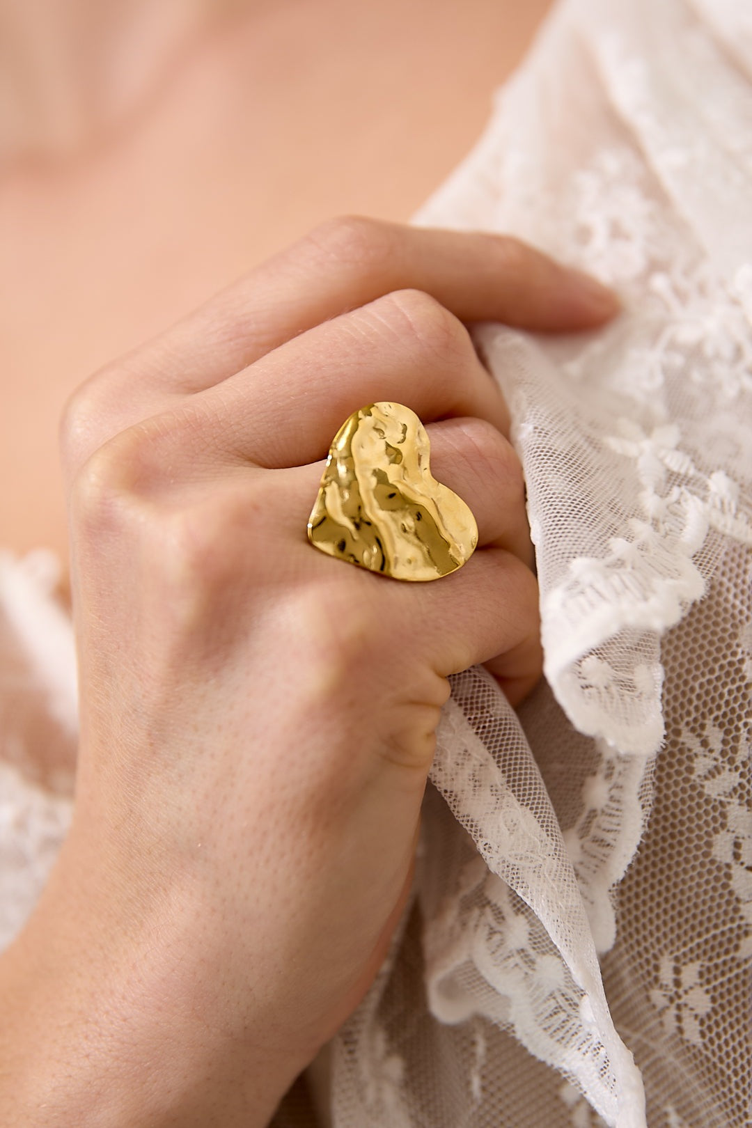 Woman's hand wearing a textured gold heart-shaped statement ring near lace fabric