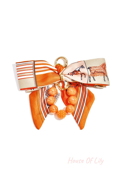 Orange patterned fabric bow with glossy matching orange and textured beads bag charm attached to gold clasp