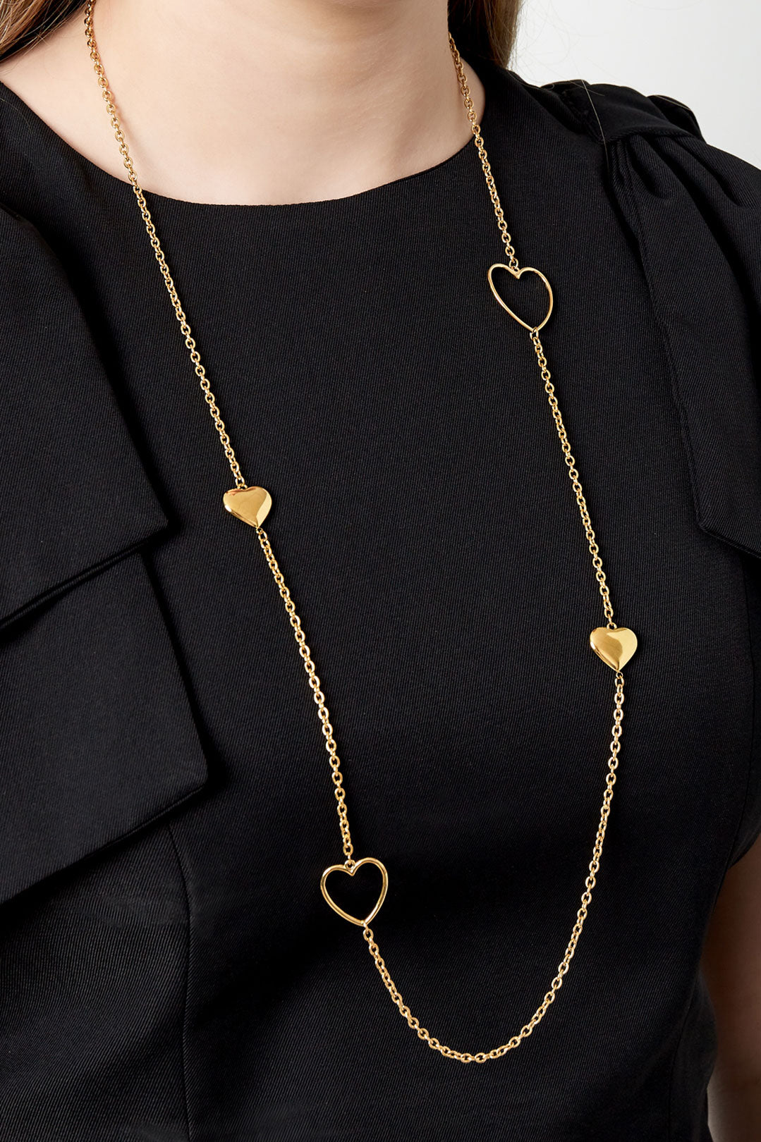 Woman wearing a long gold chain necklace with alternating solid and open heart-shaped charms over a black top