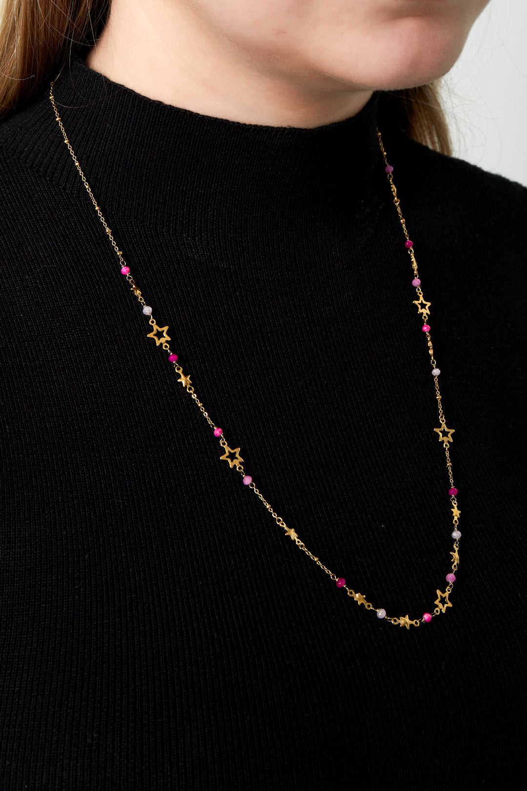 Model wearing the gold-toned star charm necklace with pink and white beads over a black ribbed turtleneck sweater