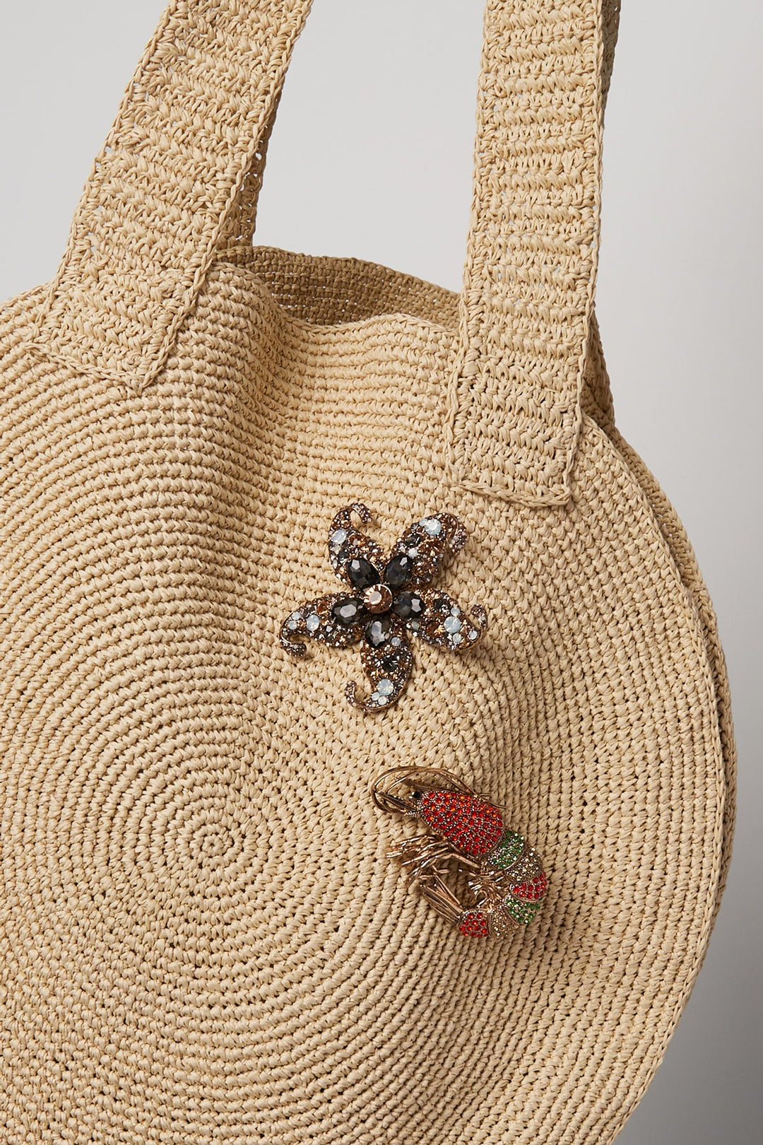 Two brooches pinned on a woven beige tote bag: a starfish-shaped floral brooch with champagne and gray rhinestones and a shrimp-shaped brooch with red and green rhinestones