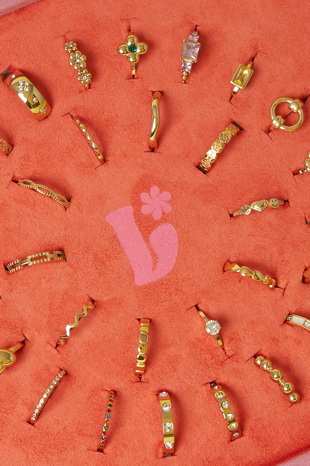 Close-up detail of gold-tone rings on coral-orange velvet display featuring rhinestones, floral and heart shapes