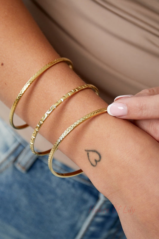 Three gold textured bangles worn stacked on wrist with a small heart tattoo and nude top
