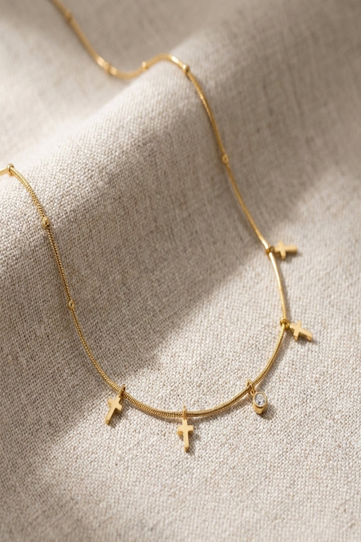 Close-up of a delicate gold cross necklace with rhinestones, resting on soft neutral fabric, emphasizing fine details and sparkle.