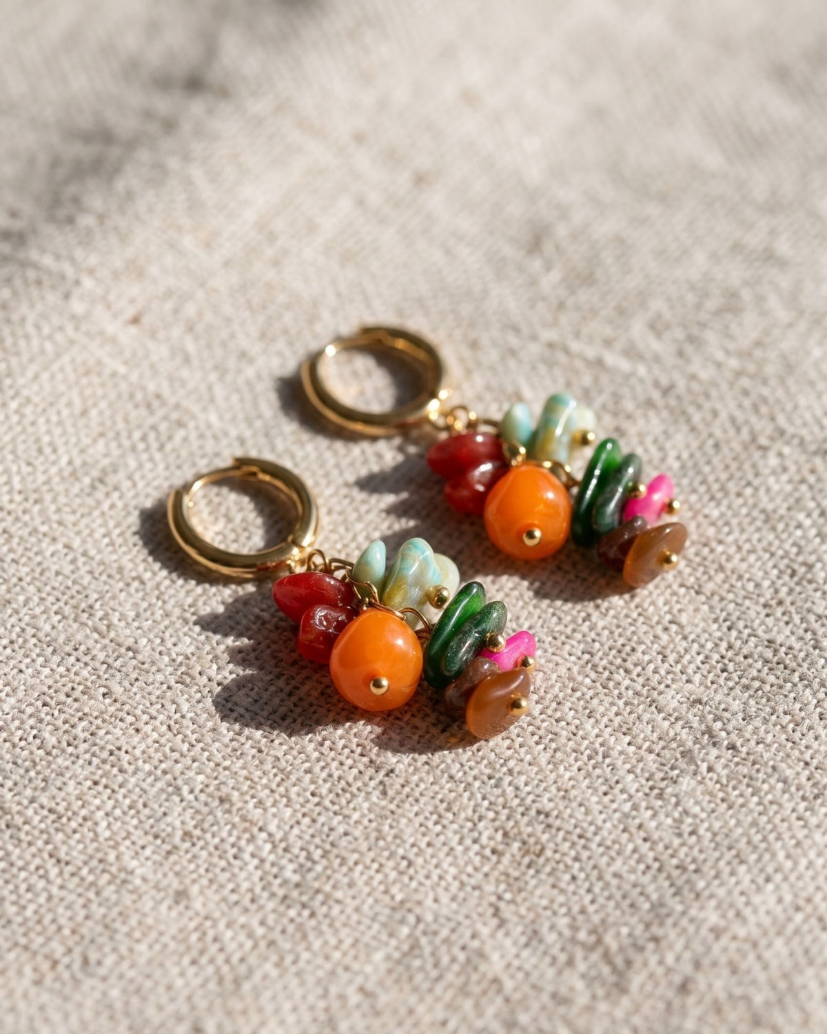 Petite stainless steel hoops with multi-colored stone beads resting on a textured linen surface.