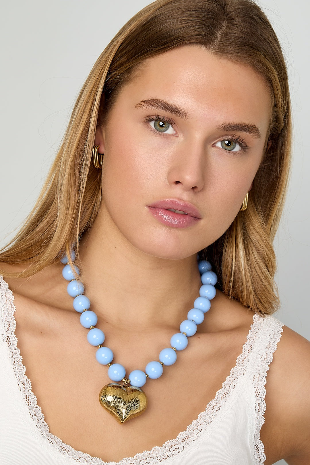 Model wearing baby blue chunky beaded necklace with gold heart pendant and white lace-trim tank top