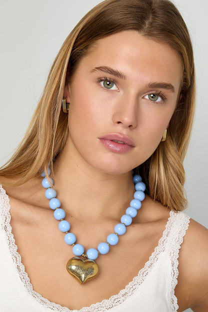 Model wearing baby blue chunky beaded necklace with gold heart pendant and white lace-trim tank top