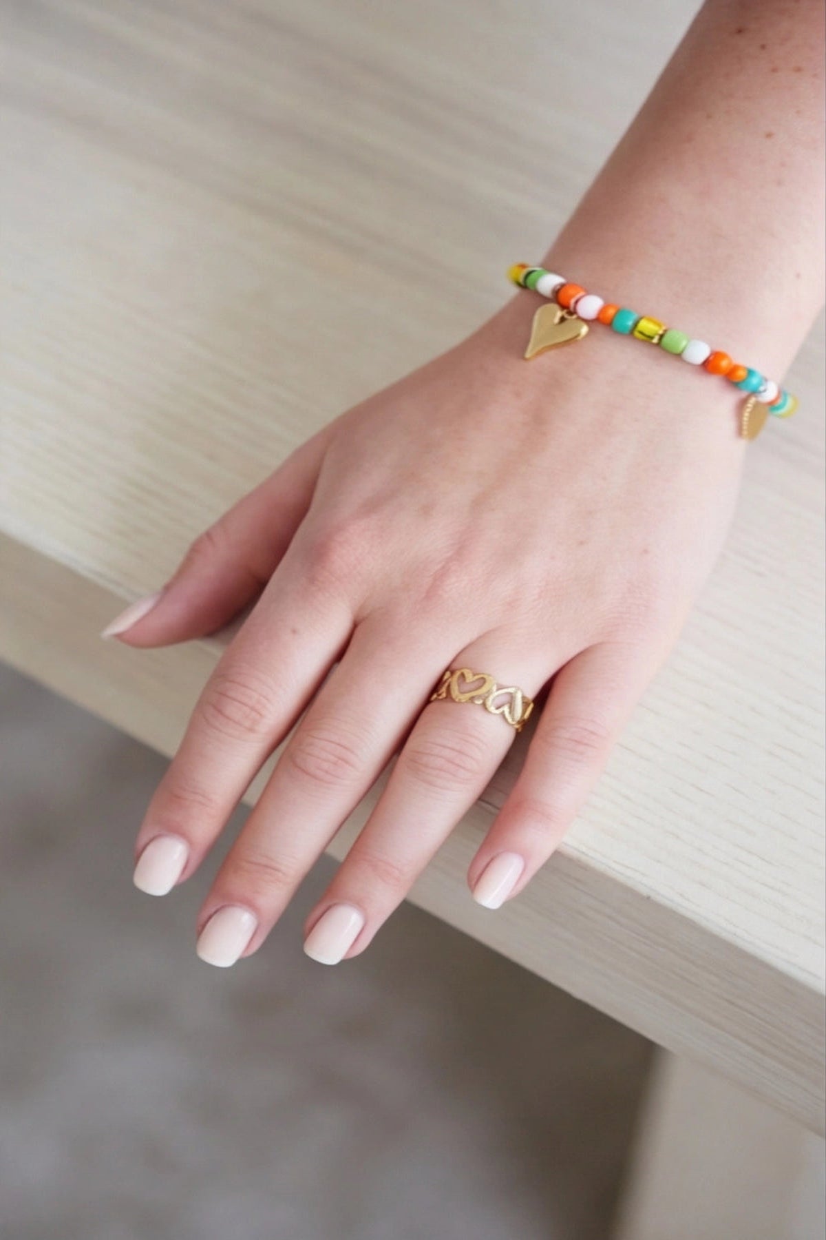 Hand wearing a gold ring and colorful beaded bracelet on a neutral background