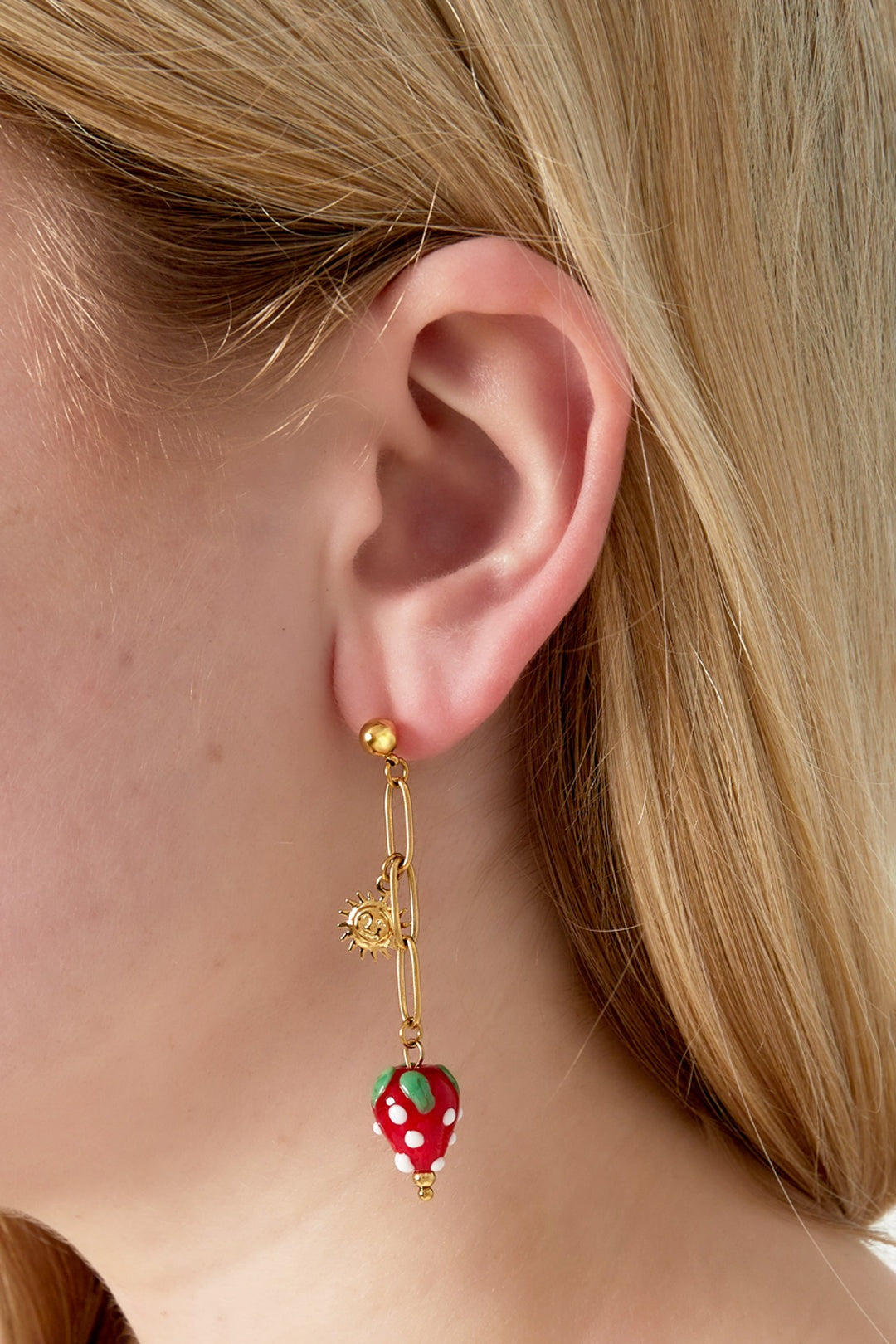 Close-up of a woman wearing gold drop earrings with sun charm and red strawberry enamel pendant