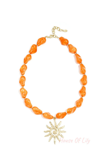 Vibrant orange acrylic beaded necklace with a textured gold sun pendant shown on a white background