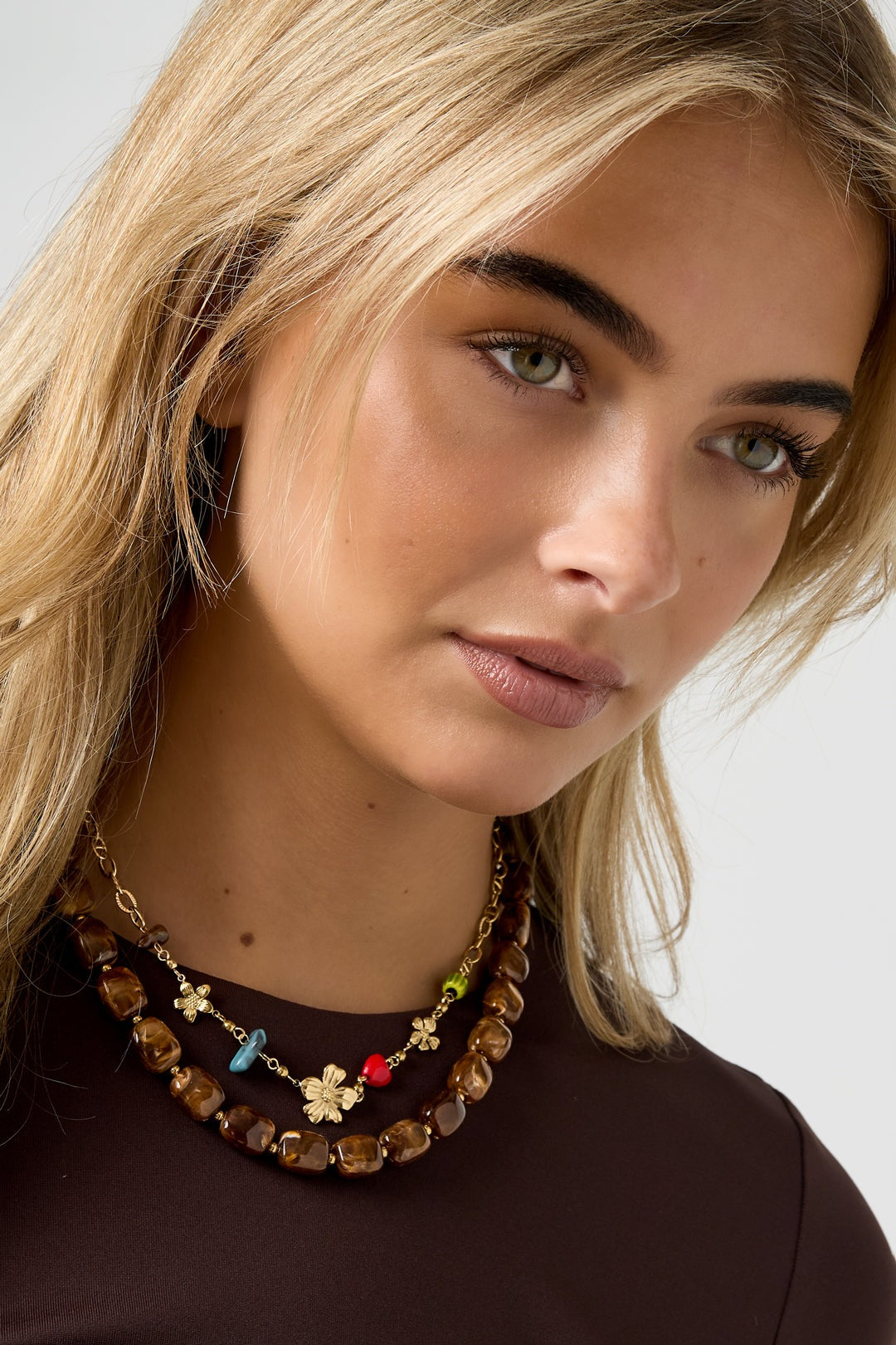 Model wearing layered necklaces including gold flower charm chain with colorful beads and chunky brown necklace