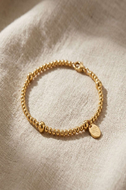 Small beaded bracelet in gold or silver resting on linen fabric, showcasing polished beads and lobster clasp detail.