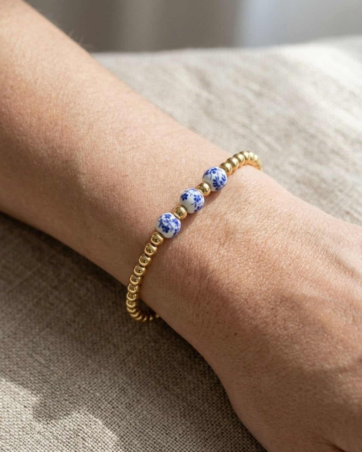 Delicate ceramic bead bracelet with blue floral patterns and gold accents worn on a wrist against a natural linen background.
