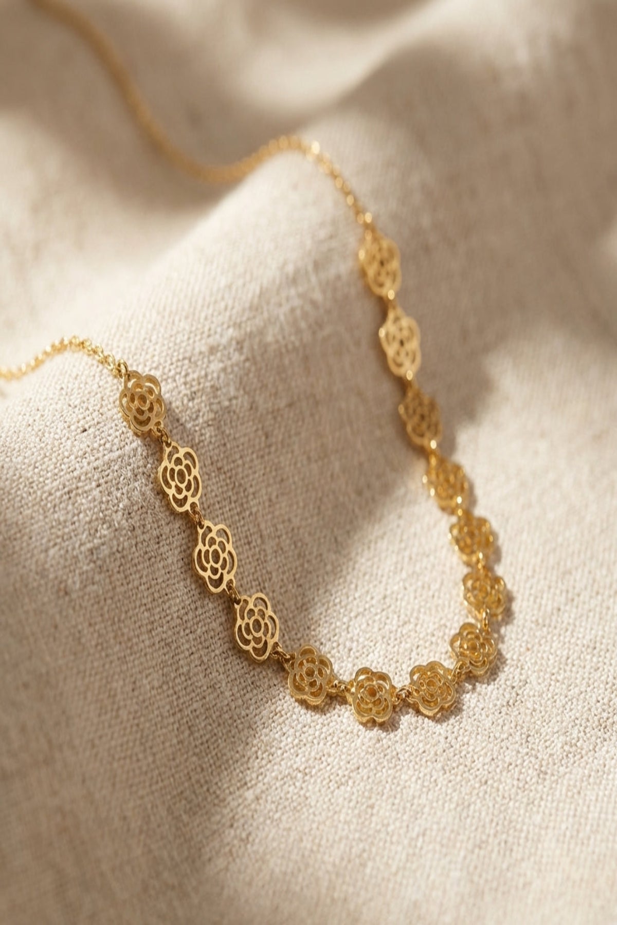 Close-up of a delicate floral chain necklace with tiny flower charms, gold-plated stainless steel, on a soft natural textured background, showcasing fine details and luster.