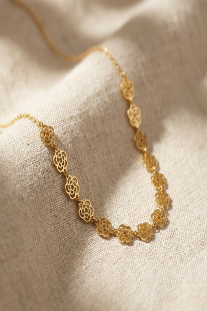 Close-up of a delicate floral chain necklace with tiny flower charms, gold-plated stainless steel, on a soft natural textured background, showcasing fine details and luster.