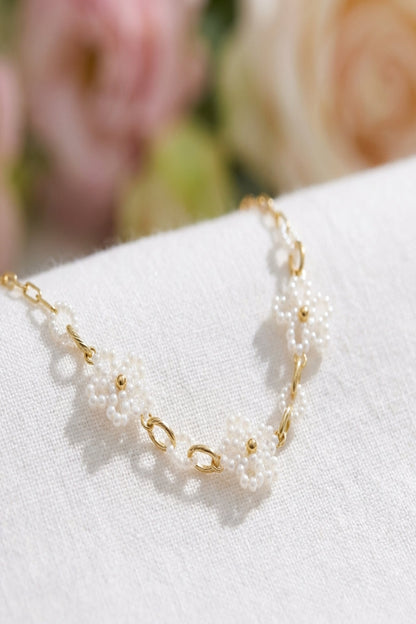 Close-up of a delicate floral pearl necklace showcasing tiny white pearls in flower shapes on a thin gold chain, with soft pastel background and natural daylight.