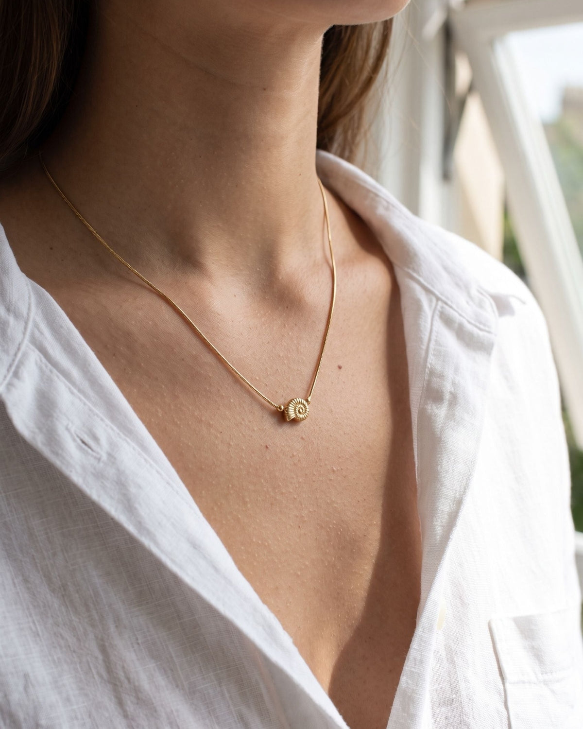 A petite gold seashell pendant on a delicate chain worn against a white linen shirt, capturing the textured spiral detail.
