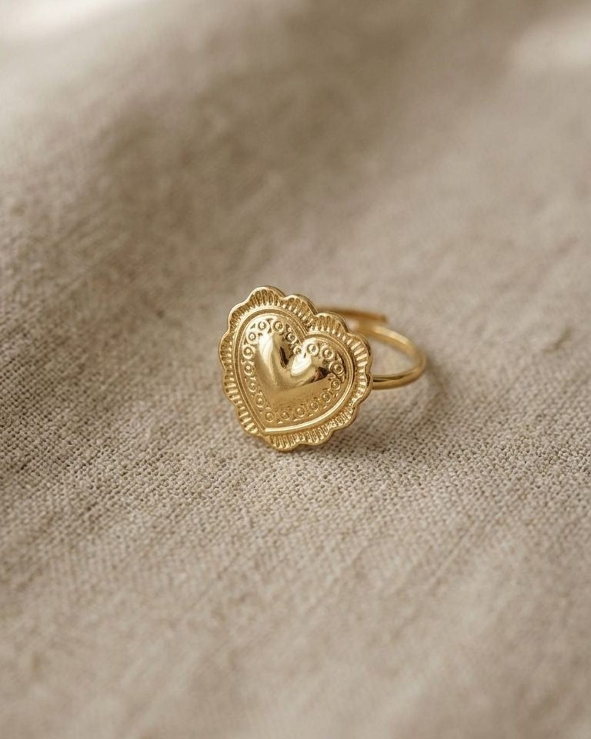 Small gold-tone hammered heart ring with scalloped edges on beige fabric, highlighting textured finish and vintage-inspired design.