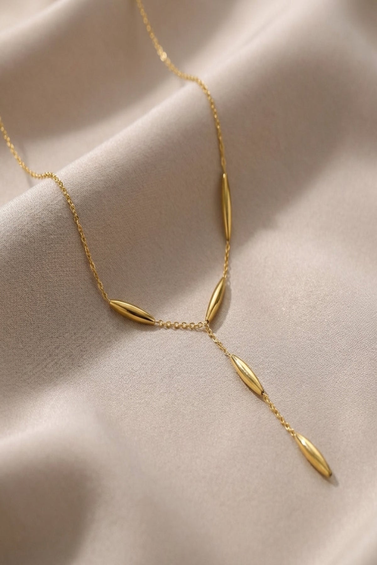 Close-up of a small, elegant gold Y-shaped necklace resting on neutral silk fabric, highlighting its fine chain and polished beads with soft natural lighting.