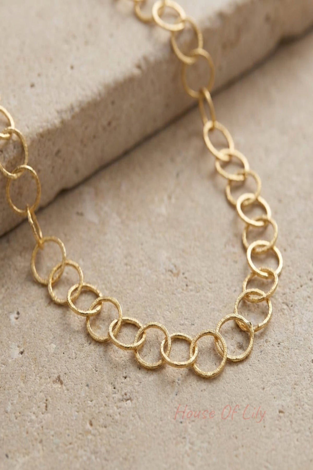 Close-up of a delicate gold-plated stainless steel loop chain necklace with fine links, showcased on a neutral textured surface with soft lighting.