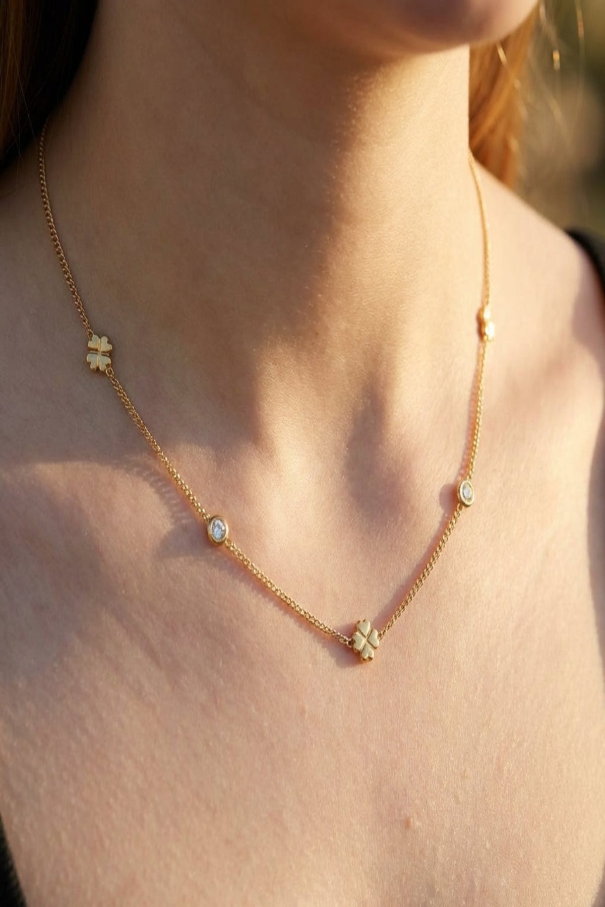High-resolution close-up of a petite gold Lucky Clover Chain Necklace worn on a female model's collarbone, featuring tiny clover charms and sparkling rhinestones in warm golden hour lighting.