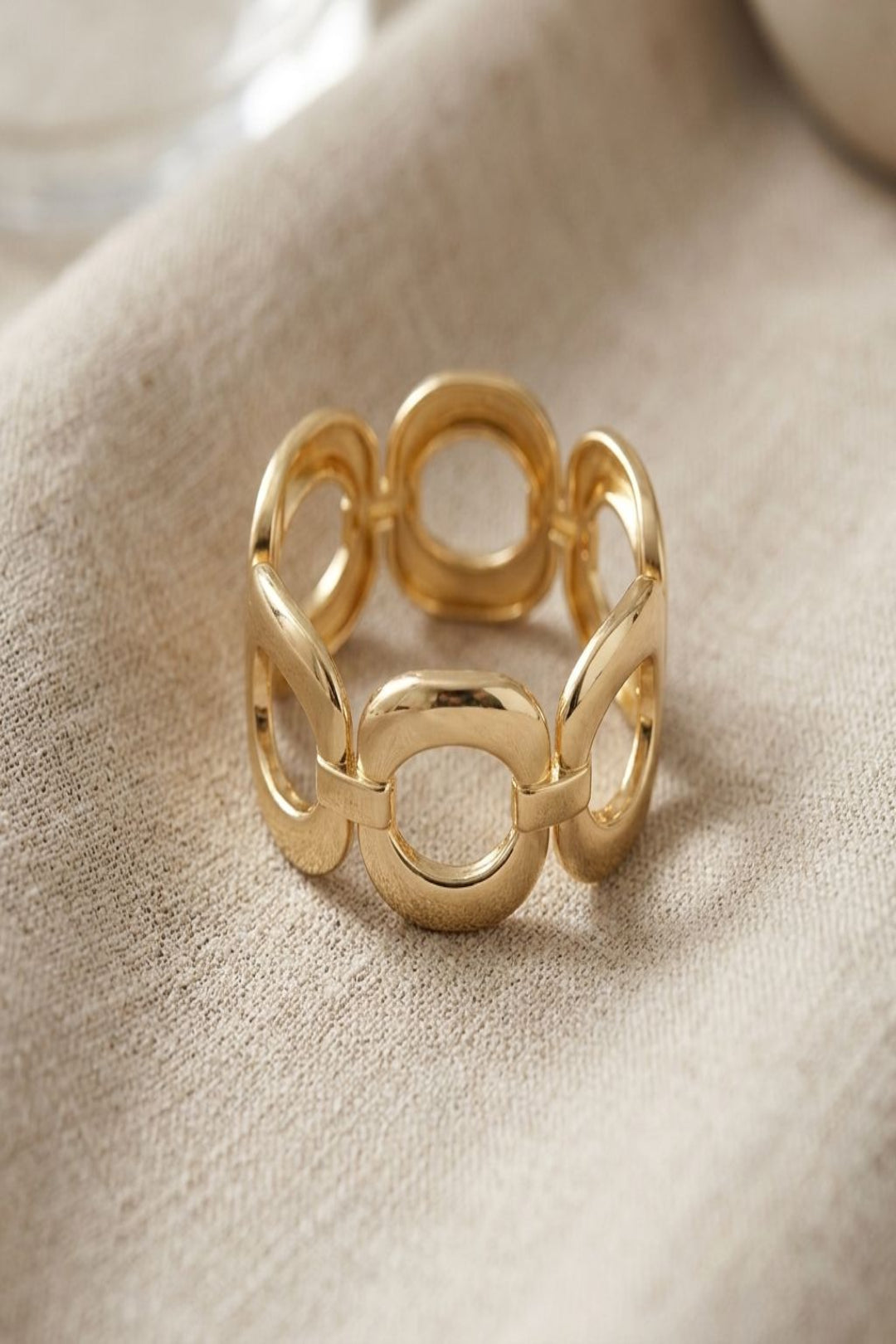 Close-up of a small oval alloy bracelet resting on beige linen, highlighting its irregular shape and subtle sheen with natural light.
