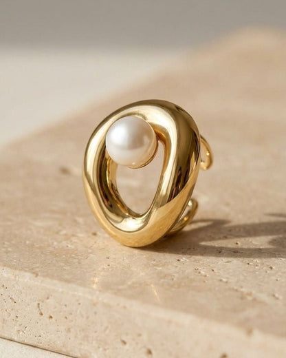 Close-up of a delicate sculpted pearl statement ring in polished brass with 18k gold plating, on a natural stone with soft sunlight highlighting its organic curves.