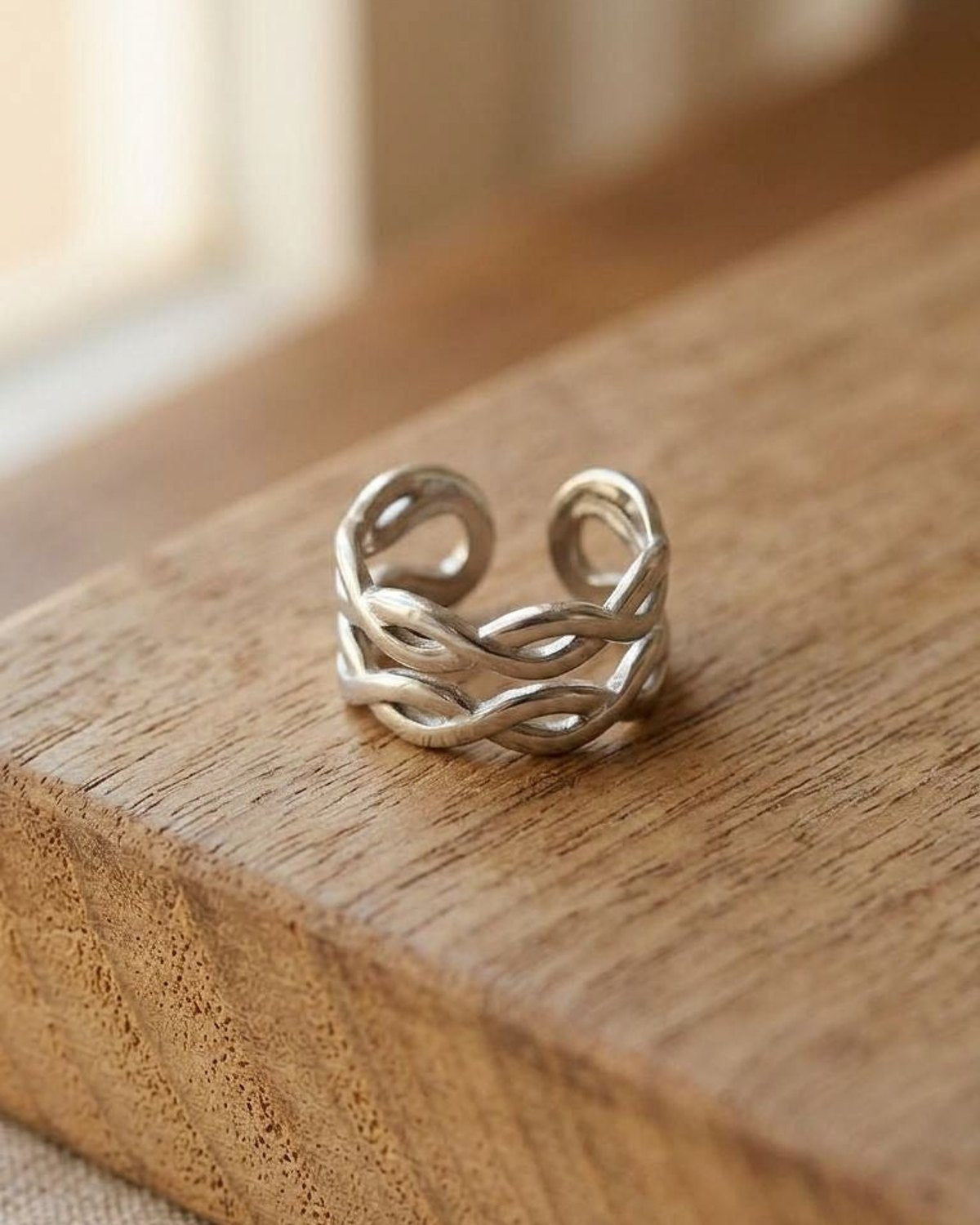 Miniature stainless steel ring with a double twist design on a wood surface, lit by natural light, highlighting its sculptural interwoven silhouette.