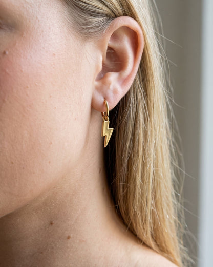 A delicate stainless steel drop earring with a subtle lightning bolt pendant worn on a blonde woman's ear.