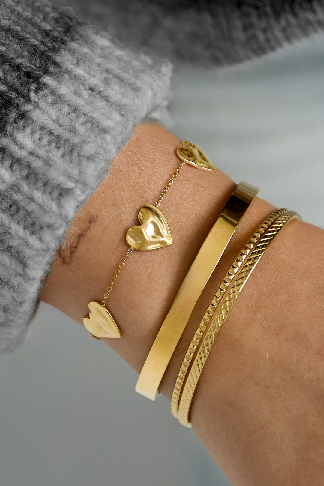 Wrist wearing gold tone charm bracelet with three sculpted hearts alongside two other gold bracelets and a gray knit sleeve