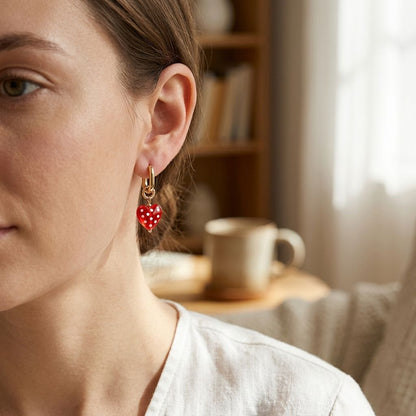 Woman wearing dotted heart drop earrings in cozy indoor setting, showcasing small, feminine jewelry with natural sunlight.