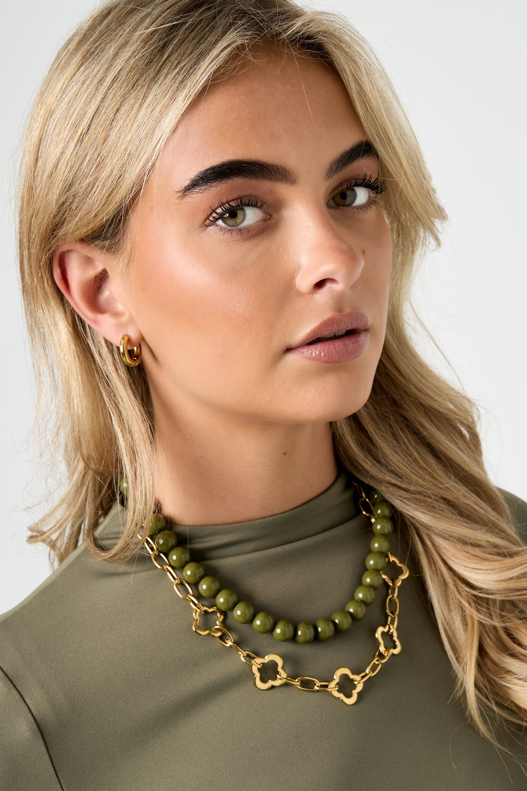 Model wearing a gold-tone clover link chain necklace layered with an olive green bead necklace, paired with gold hoop earrings