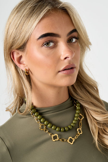 Model wearing a gold-tone clover link chain necklace layered with an olive green bead necklace, paired with gold hoop earrings