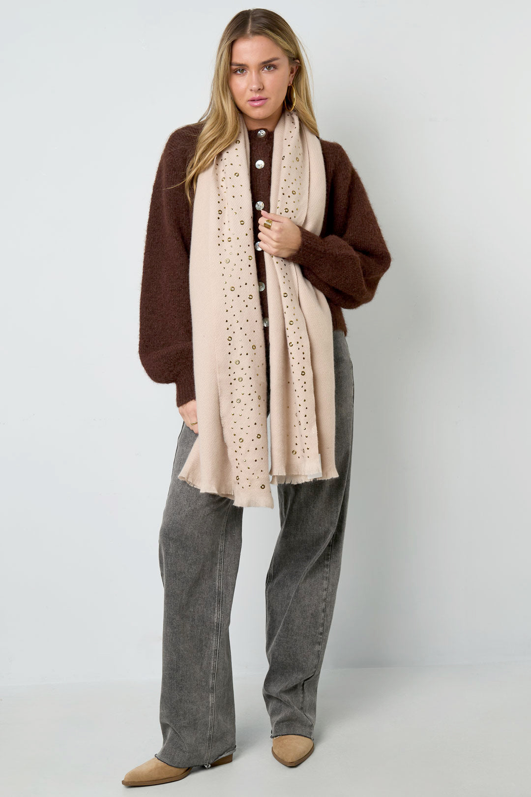 Woman wearing cream studded scarf draped over dark brown sweater and gray pants posing front view