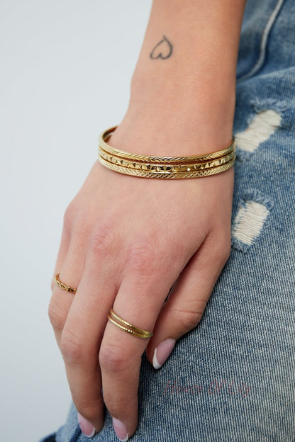 Woman's hand resting on denim-clad leg, wearing a set of three gold-tone slim bangles and matching rings