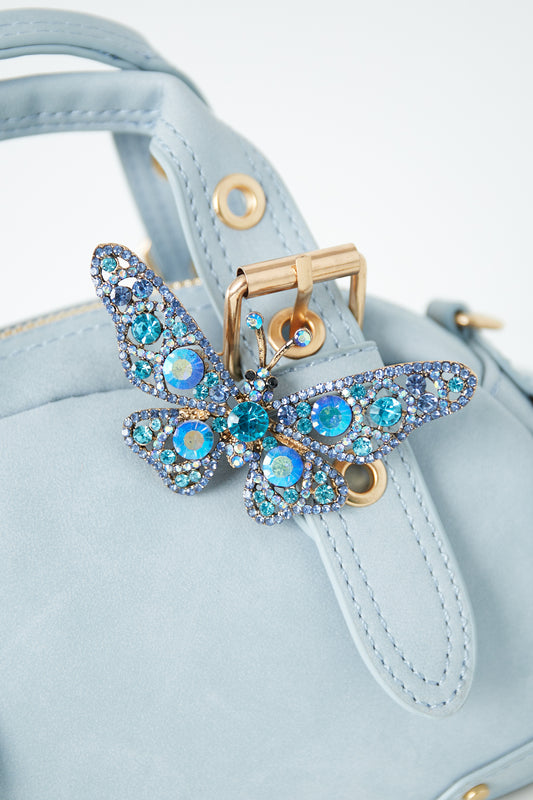 Blue rhinestone butterfly brooch pinned to a light blue handbag strap with gold buckle hardware