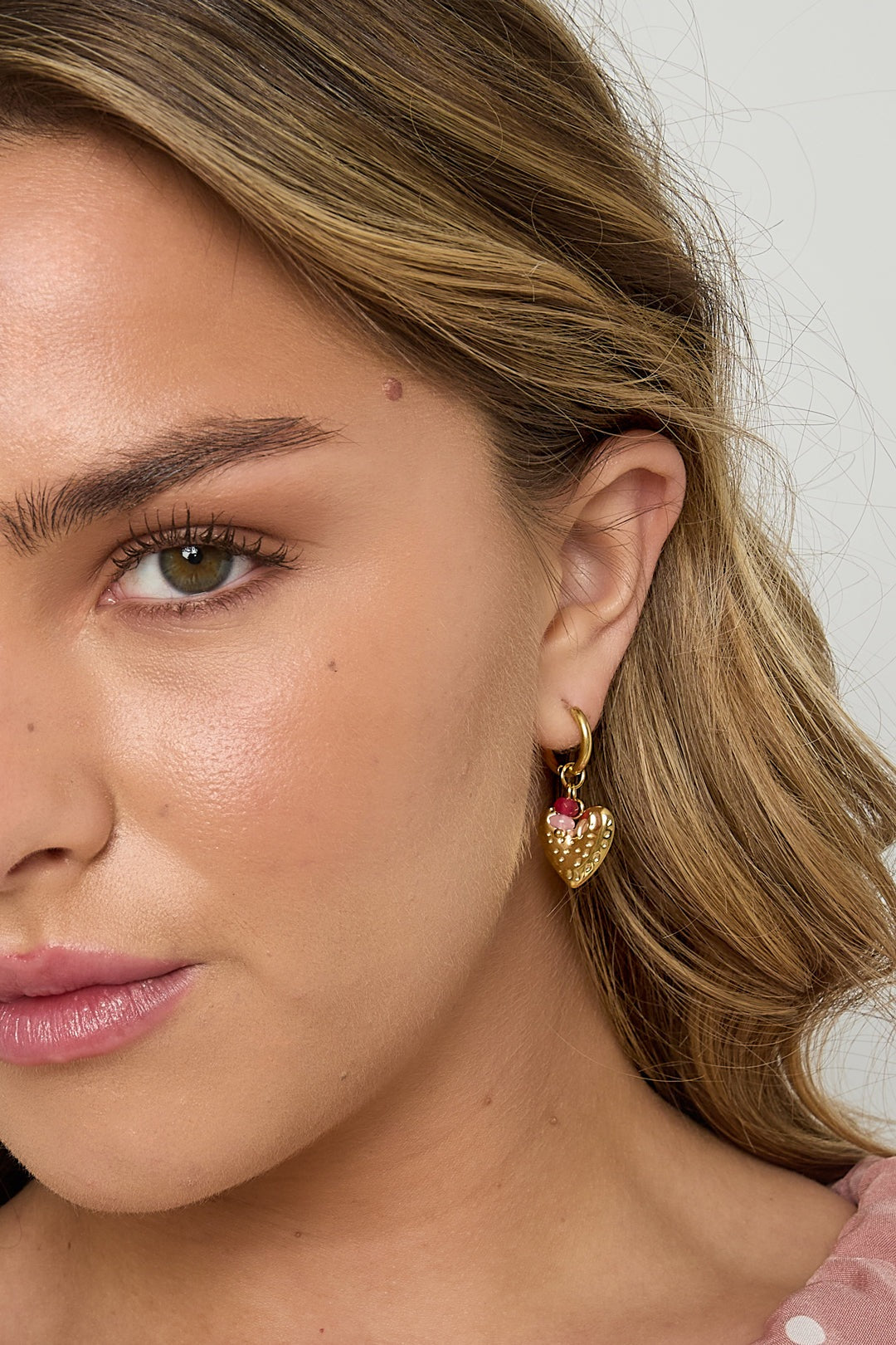 Close-up of a woman wearing one gold heart drop earring with pink and red beads, highlighting the playful design