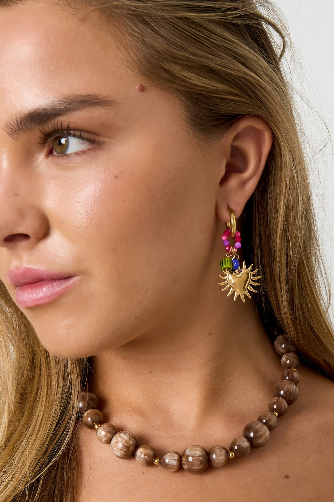Close-up of a woman wearing gold sunburst heart hoop earrings with colorful beads, showcasing youthful style