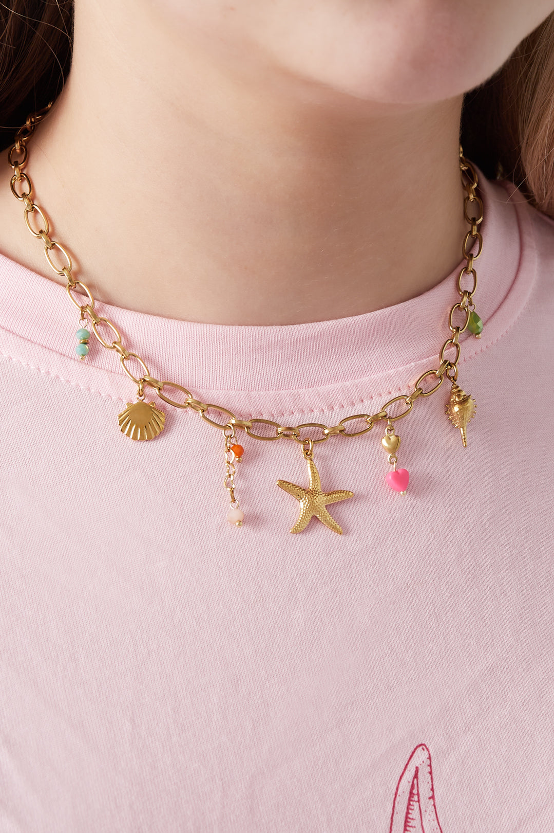 Close-up of gold-tone charm necklace worn by woman with pastel pink top, showing beach-themed charms