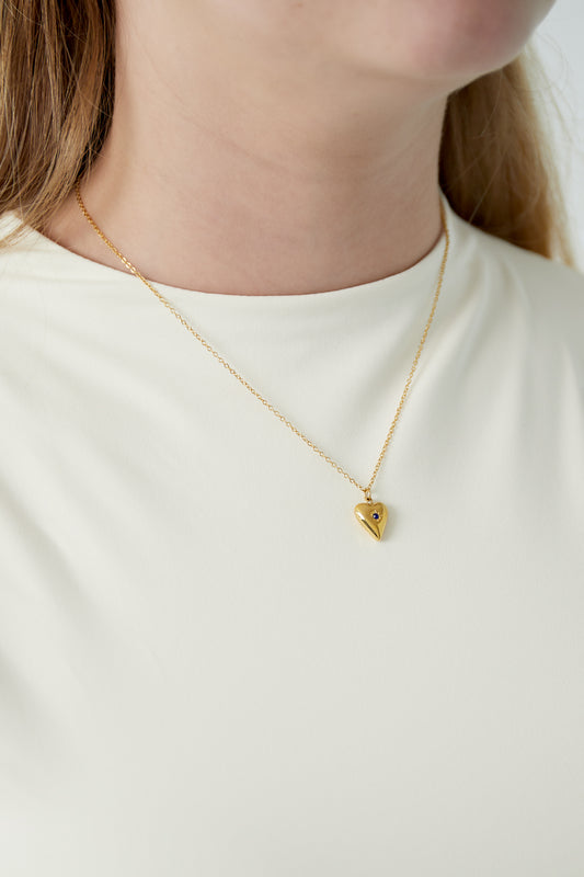 Close-up of model wearing gold heart necklace with dark blue birthstone on white top