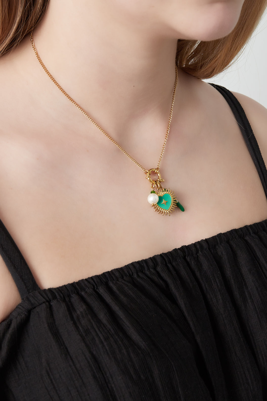 Close-up of necklace with green heart and pearl charms worn by a woman in a black top against white background