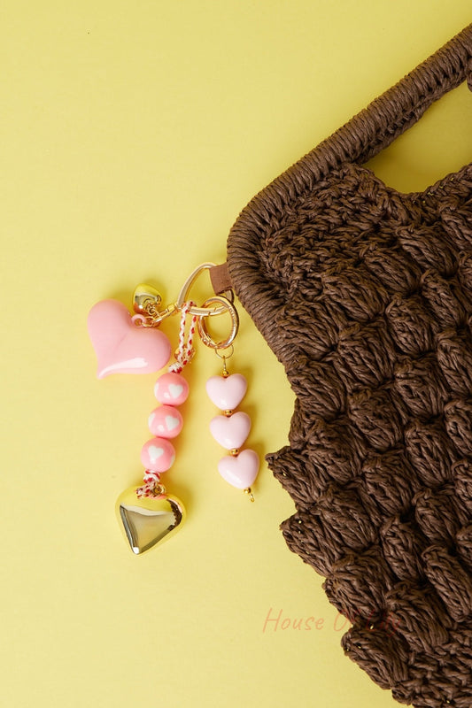 Pink-themed bag charms attached to woven brown bag on yellow background, featuring heart pendants and beads